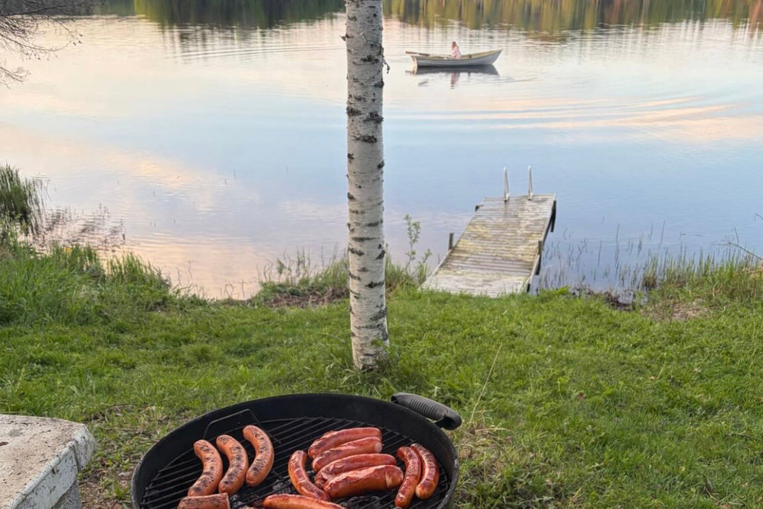 grilli järvenrannassa, jossa laituri ja nainen soutaa veneellä